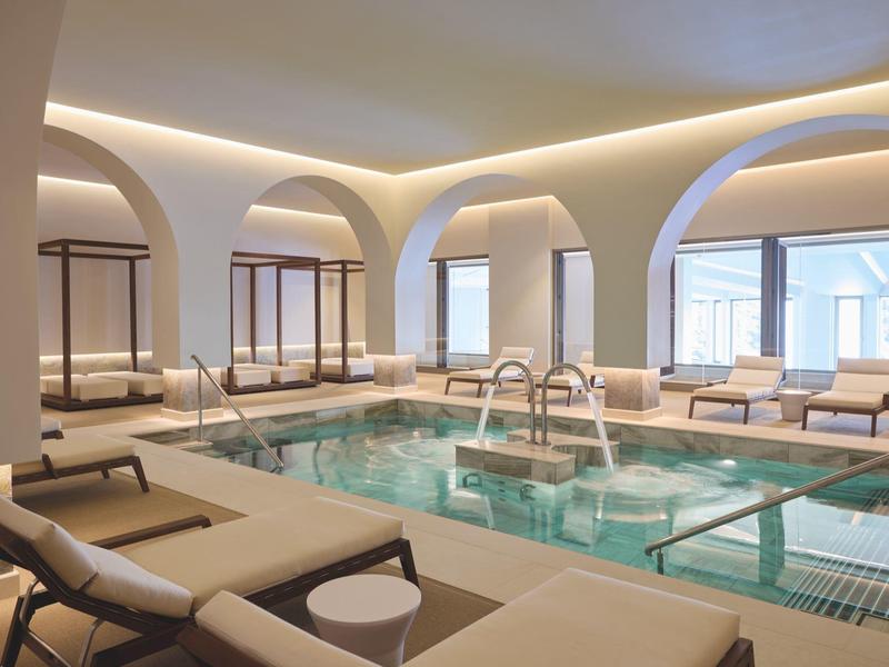 Moderní krytý bazén s bílými oblouky, lehátky a relaxační atmosférou v hotelu.