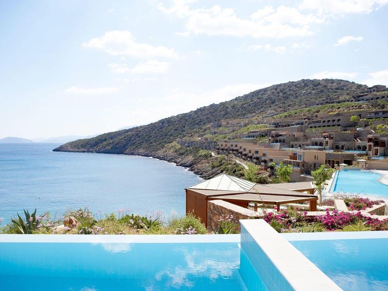 Pohled na pobřeží s bazénem a kopci s domy v pozadí.