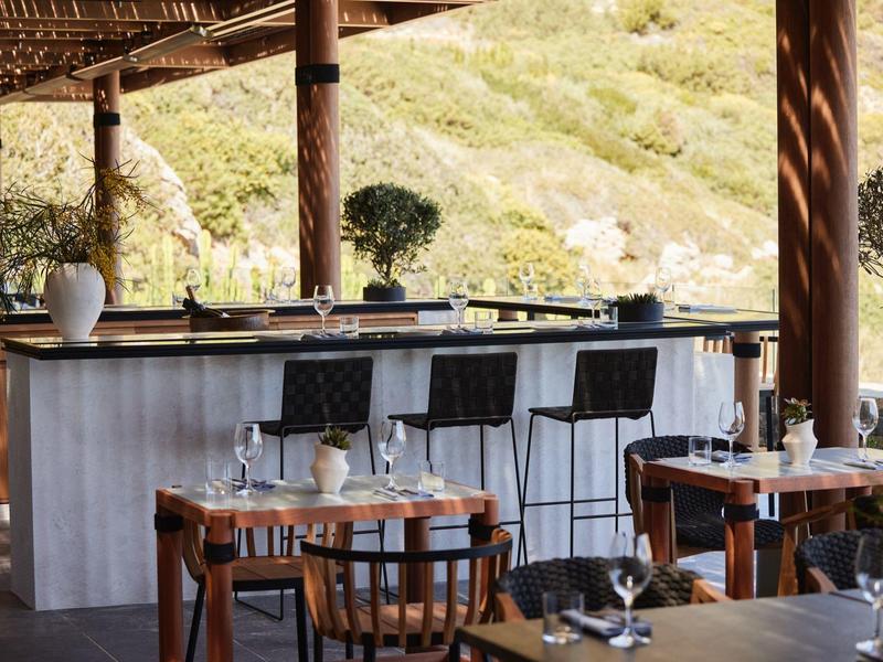 Moderní venkovní bar s dřevěným nábytkem a židlemi před zelenými kopci