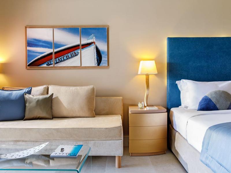 Pohodlný hotelový pokoj se šedou pohovkou, modrou postelí a moderním uměním na zdi.