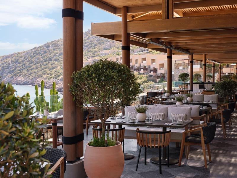 Venkovní terasa s stolky a židlemi s výhledem na moře a hory pod jasnou oblohou.