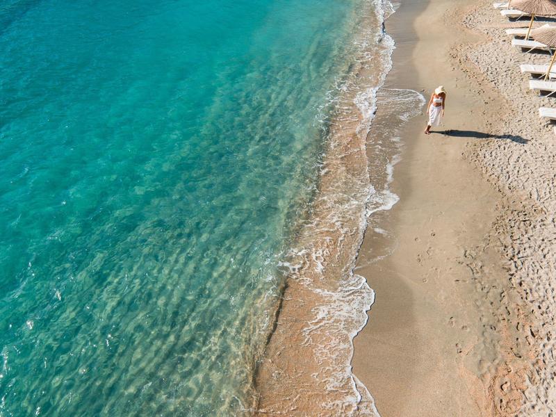 Čistá modrá voda se setkává s písčitou pláží s roztroušenými lehátky a jednou osobou.