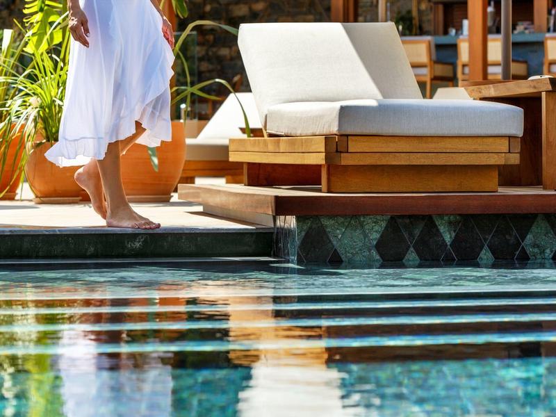 Osoba v bílém županu kráčí vedle bazénu vedle pohodlného lehátka.