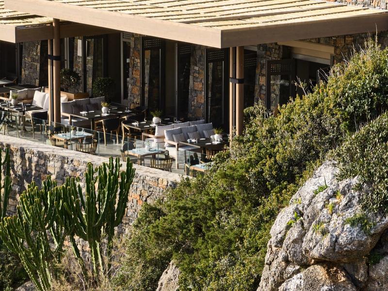 Hotelová terasa s stoly a židlemi, obklopená skalami a vegetací.