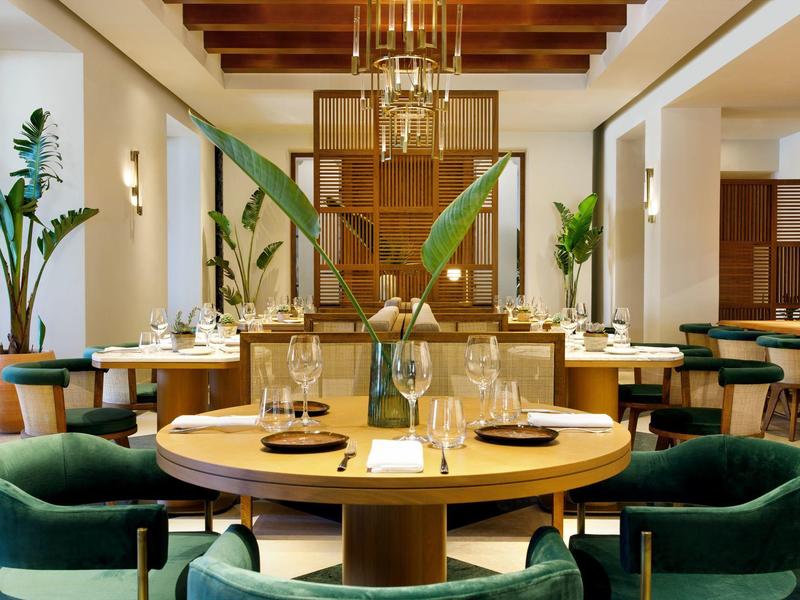 Moderní restaurace s kulatými stoly, zelenými sametovými židlemi a rostlinami ve stylovém prostředí.