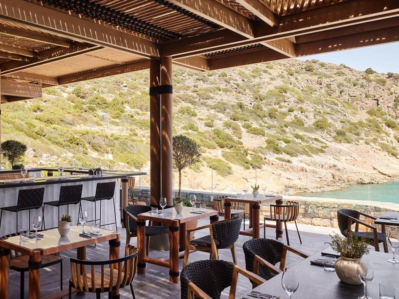 Otevřená restaurace s dřevěnými stoly a židlemi s výhledem na moře a kopce.