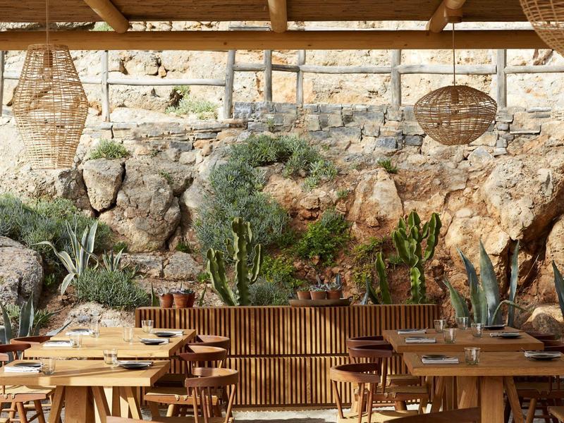 Pohodlná venkovní část restaurace s dřevěným nábytkem, rostlinami a kamennou zdí.