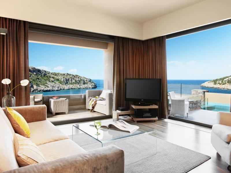 Světý obývací pokoj s velkými okny s výhledem na moře a terasu pod modrou oblohou.