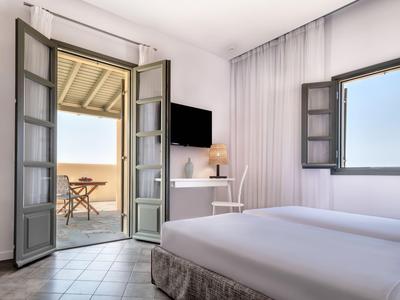 Habitación de hotel luminosa con cama doble, puerta abierta a balcón con vista al mar y ventana con luz natural.