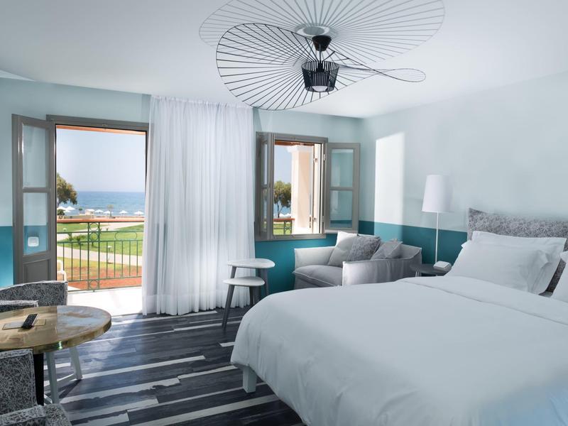 Habitación de hotel luminosa con vista al mar, cama blanca, sofá, mesa y ventilador en el techo.