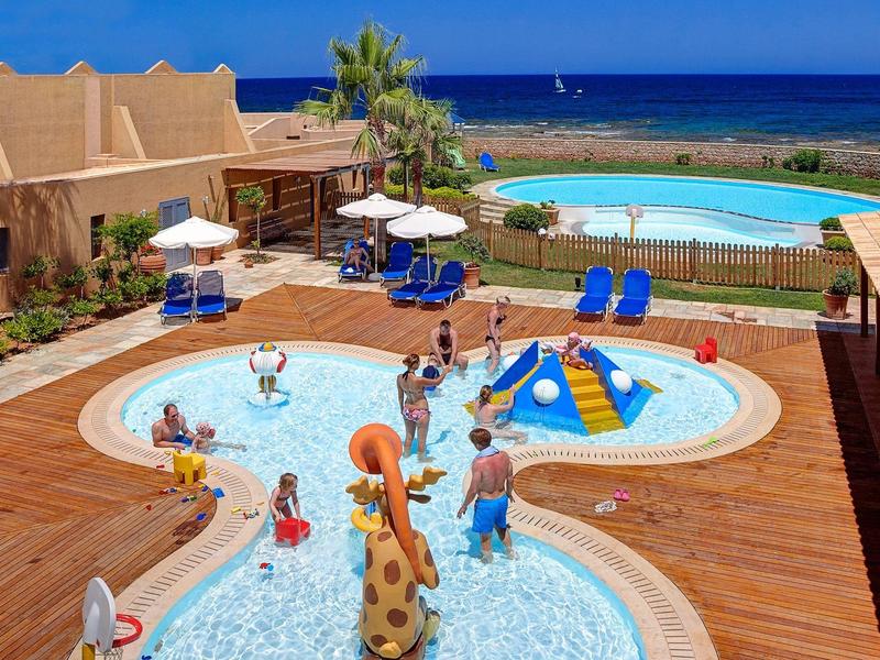 Área de piscina al aire libre con piscina para niños, juegos acuáticos y vista al mar de fondo.