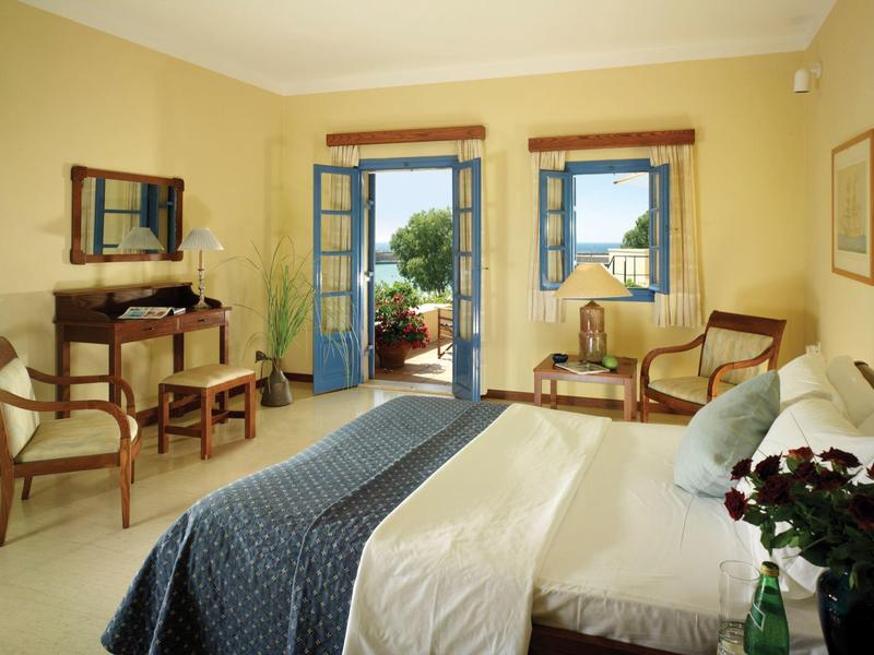 Dormitorio luminoso con cama doble, sillas de madera y balcón abierto con vistas verdes.