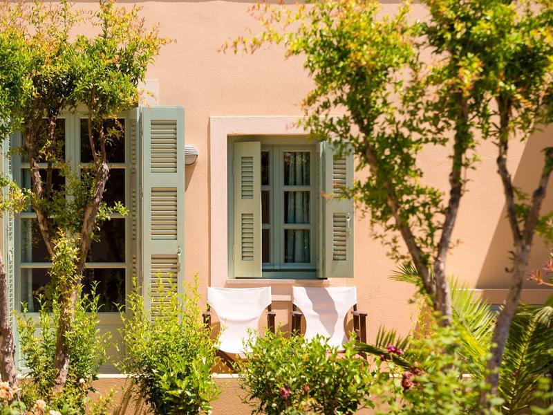 Fachada de una casa con contraventanas verdes y arbustos en primer plano bajo la luz del sol.