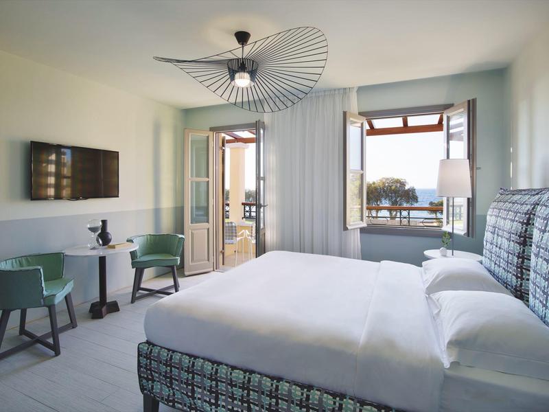 Habitación de hotel luminosa con una cama grande, dos sillas, mesa y ventana abierta con vista al mar.