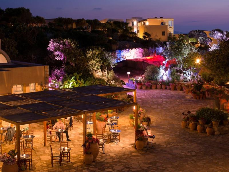 Vista nocturna exterior de un restaurante iluminado con mesas y sillas en un complejo hotelero.