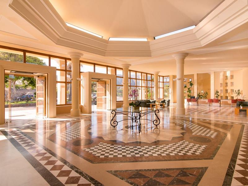 Amplio vestíbulo de hotel con suelo de mármol y mesas bajo un alto y luminoso techo abovedado.