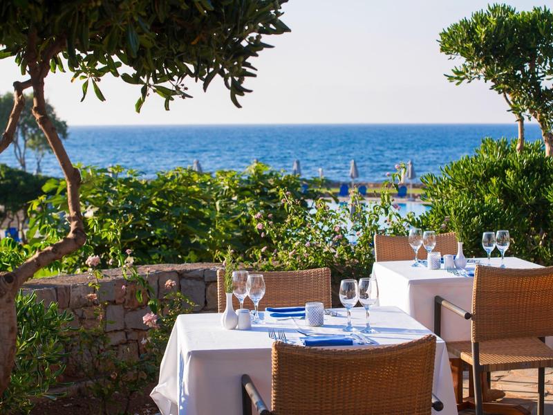 Terraza con mesas servidas y vista al mar, rodeada de árboles y plantas.