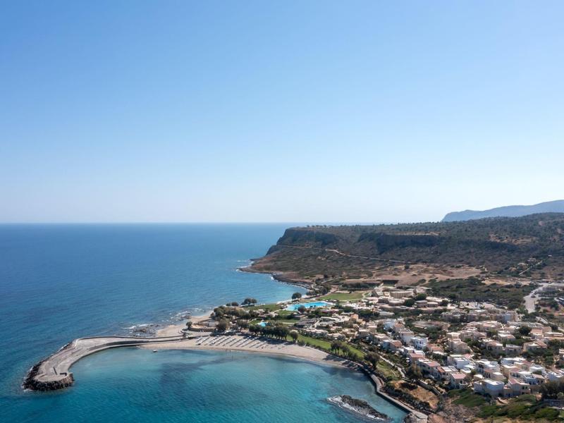 Costa con puerto, agua azul clara y casas en un paisaje soleado.