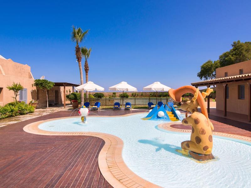 Piscina infantil con figuras divertidas y sombrillas en el jardín de un hotel bajo cielo azul.