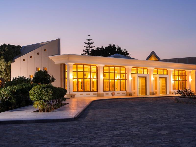 Un edificio de hotel moderno iluminado con grandes ventanas y vegetación circundante al atardecer.