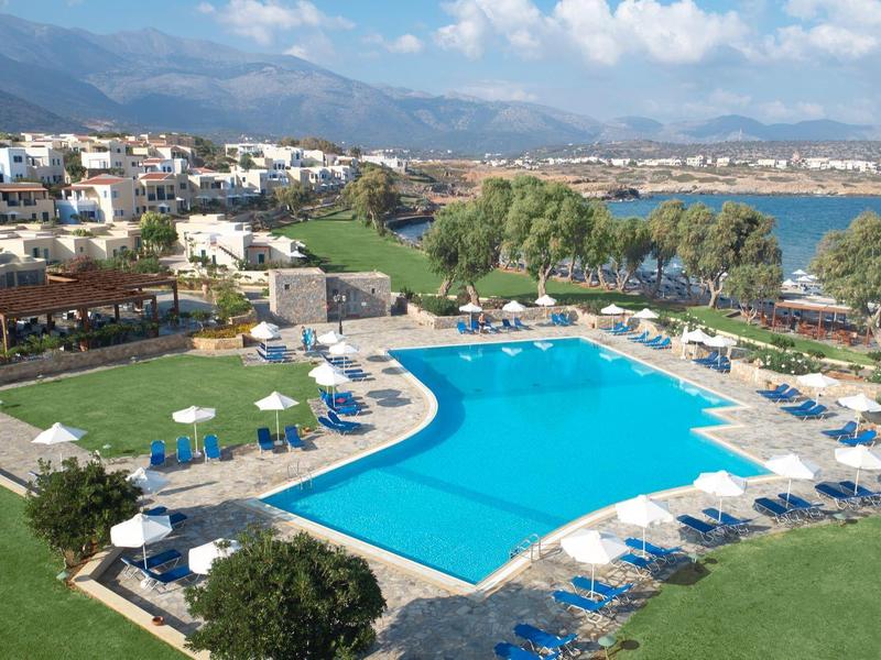 Gran piscina de hotel con tumbonas azules y sombrillas blancas, rodeada de césped verde y montañas al fondo.