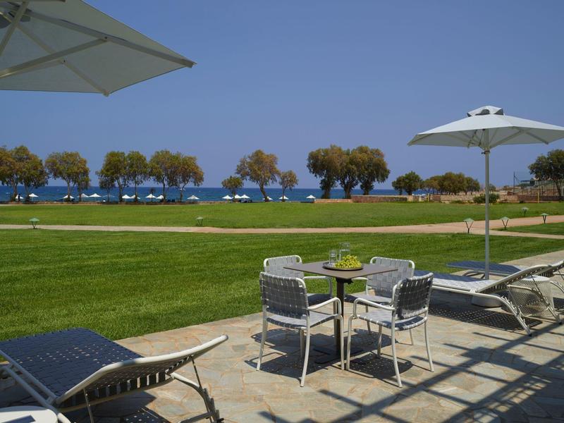 Vista de una terraza soleada con tumbonas y mesas, detrás césped y árboles junto al mar.