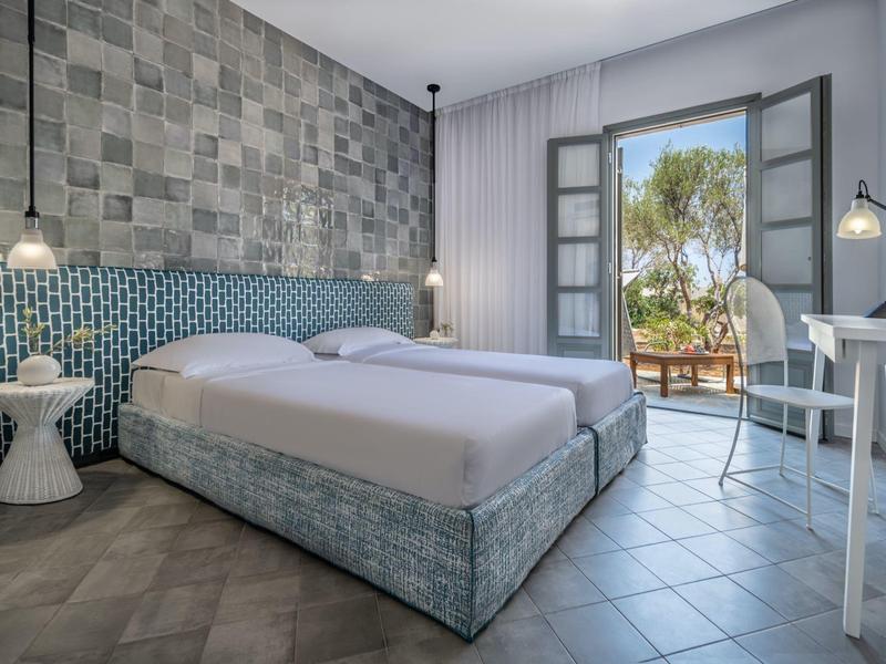 Habitación de hotel luminosa con cama grande, ventanas abiertas y decoración moderna con suelo de baldosas.