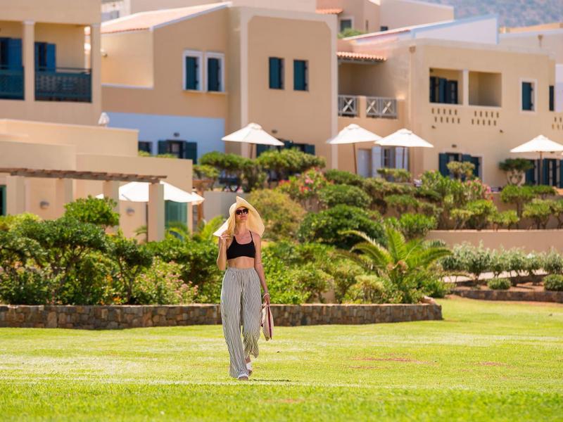Mujer con sombrero caminando sobre césped verde frente a un hotel con terrazas y sombrillas.
