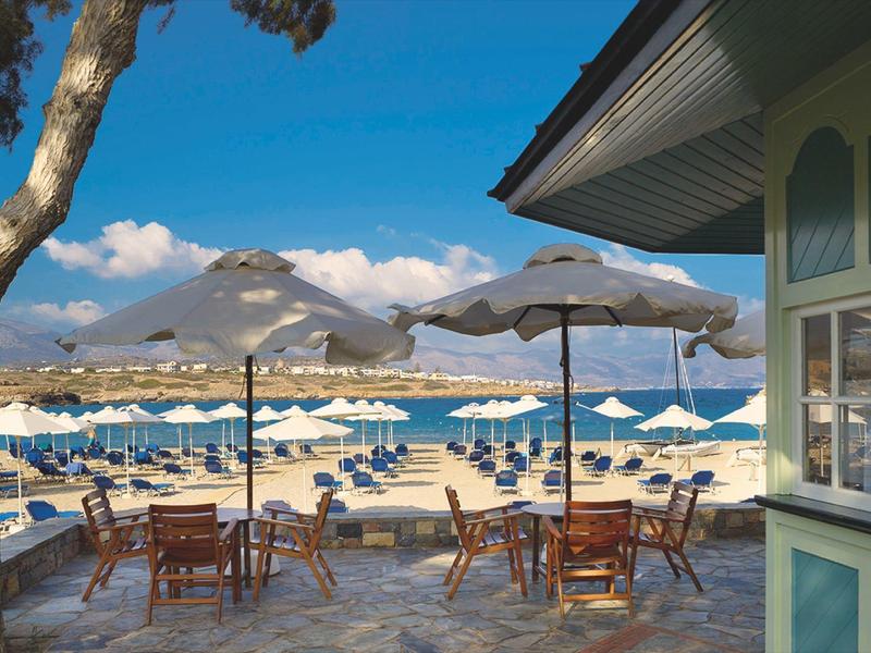Café en la playa con mesas, sombrillas y tumbonas frente al mar azul y montañas.