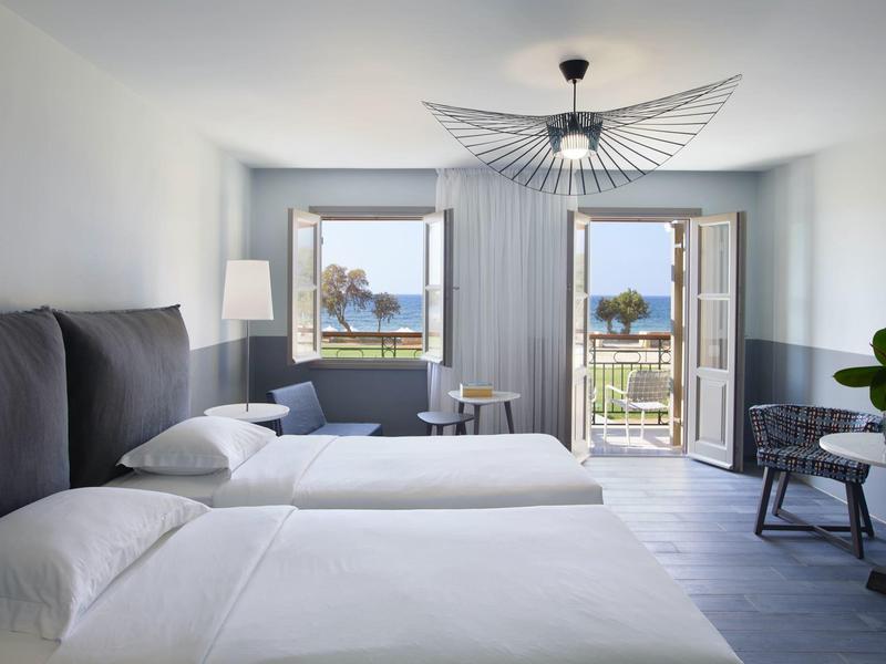 Habitación de hotel luminosa con dos camas y vista al mar a través de ventanas y balcón abiertos.