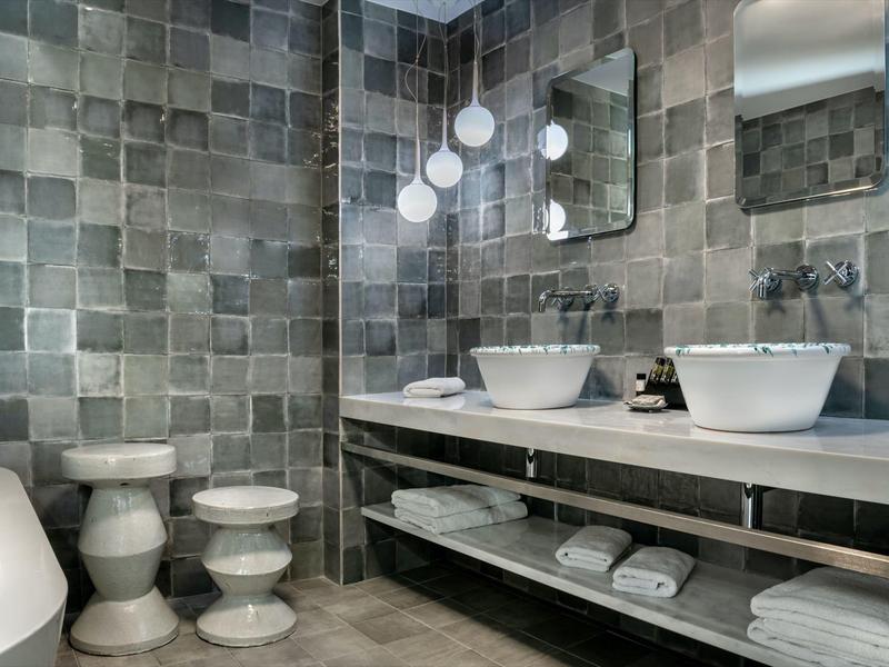 Baño moderno con azulejos grises, dos lavabos blancos y lámparas redondas en la pared.