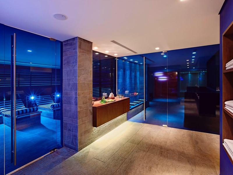 Área moderna de hotel con paredes de vidrio, armarios y zona de estar en colores neutros por la noche.