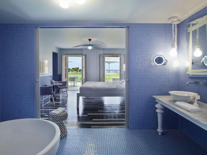 Baño azul moderno con bañera, lavabo y vista a una habitación con cama y terraza al fondo.