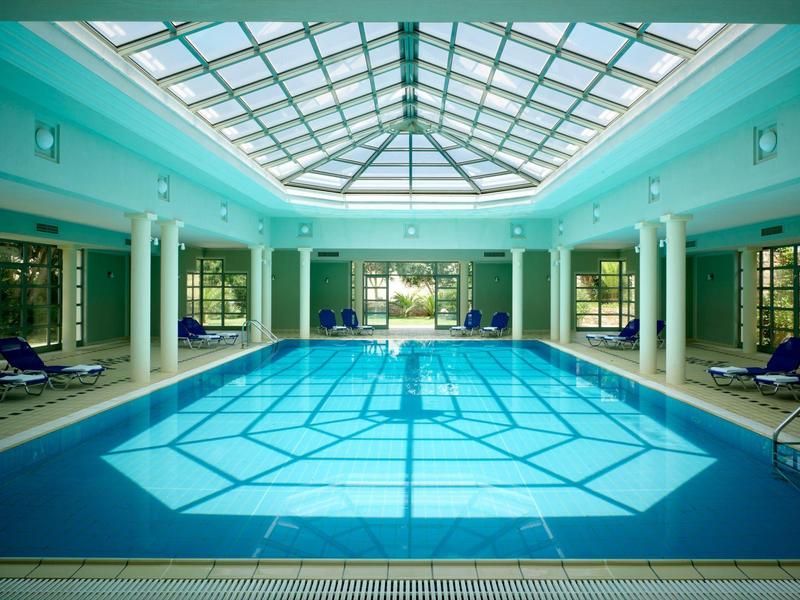 Piscina cubierta climatizada con techo de cristal y tumbonas, rodeada de grandes ventanas.