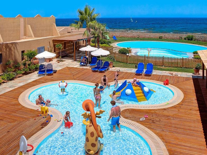 Niños chapoteando en piscina infantil junto a piscina grande con vista al mar y sombrillas.
