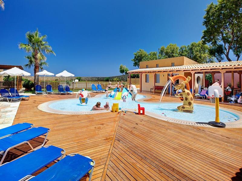 Piscina para niños con toboganes y duchas, rodeada de tumbonas azules junto a un hotel bajo el sol.