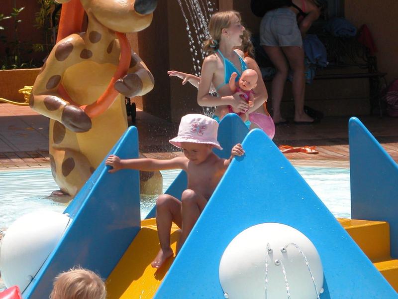 Niños juegan en un área de juegos acuáticos con una estatua grande y colorida de jirafa que rocía agua.