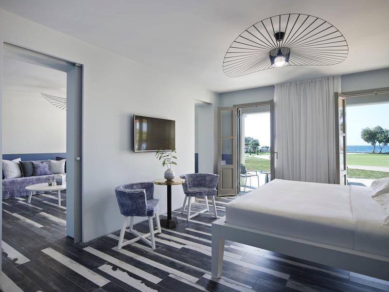 Habitación de hotel moderna con cama doble, sillones, televisión y vista al agua a través de grandes ventanas.