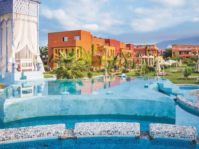 Kompleks hotelowy z niebieskim basenem, kolorowymi budynkami i zielonymi palmami pod pochmurnym niebem.