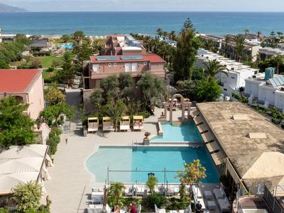 Widok na hotel z basenem, palmami i morzem w tle.