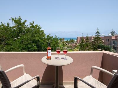 Balkon z dwoma krzesłami i małym stolikiem z napojami, widok na drzewa i morze w tle.