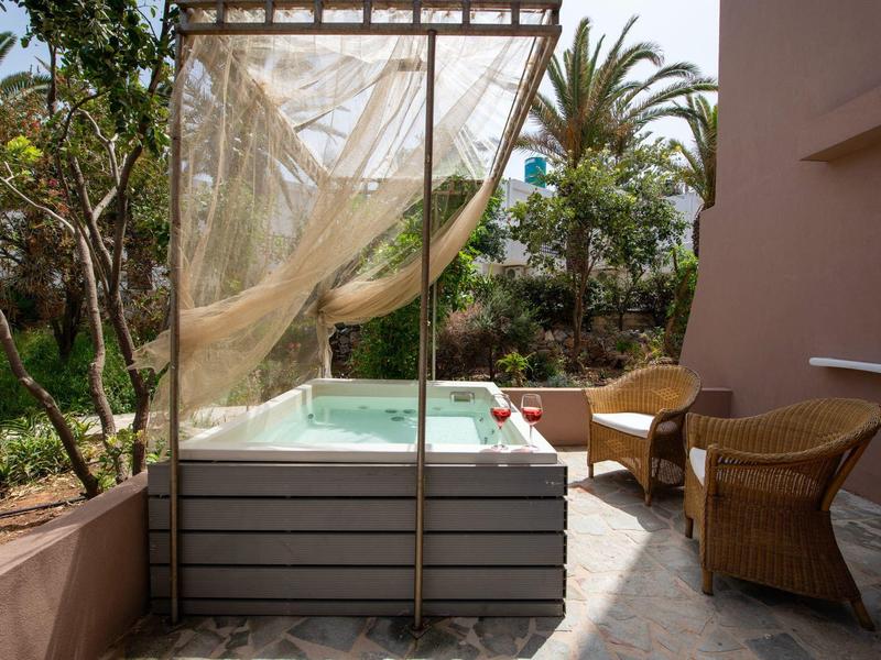 Jacuzzi na tarasie z widokiem na ogród, obok dwa wiklinowe krzesła i zasłona.