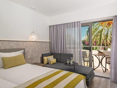 Modern hotel room with bed, gray sofa, and balcony overlooking palm trees.