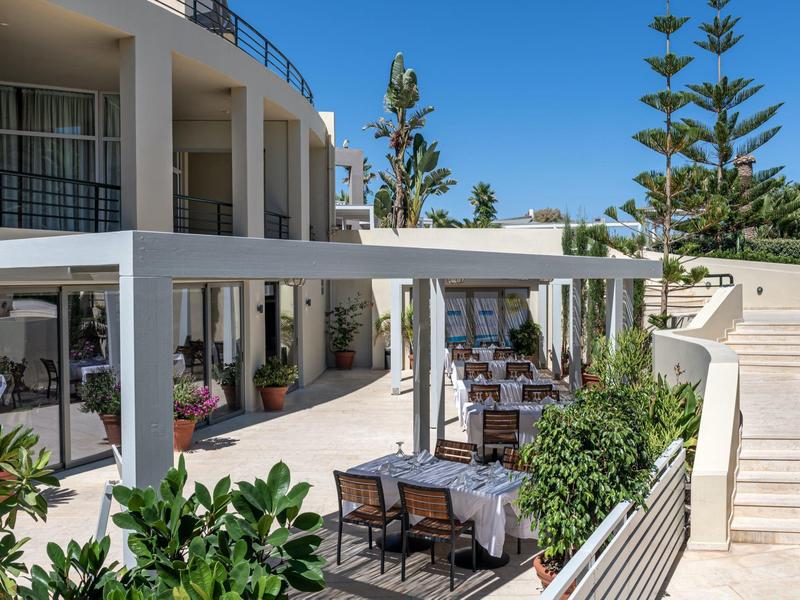 Outdoor hotel seating area with tables, chairs, umbrellas, and greenery under a blue sky.