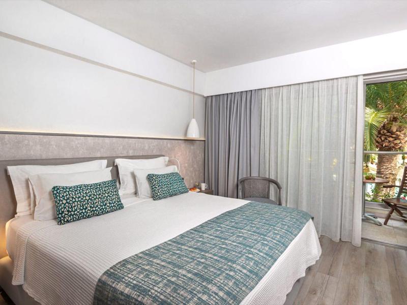 Bright hotel room with large bed, teal accents, wooden floor, and terrace with greenery.
