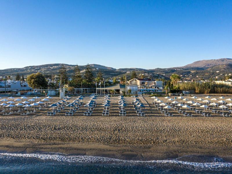 Beach with rows of sunbeds and umbrellas under clear blue sky by calm sea and hills.
