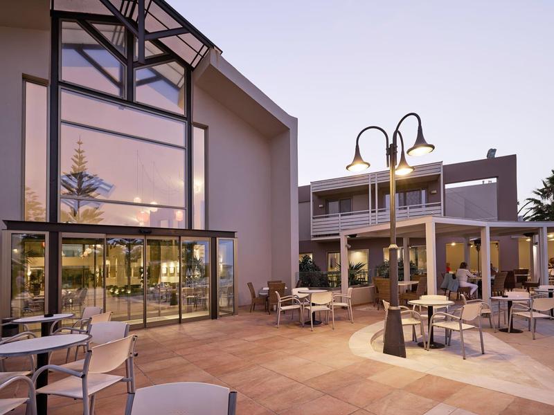Outdoor seating area of a modern hotel restaurant at dusk with glass walls and patio lamps.