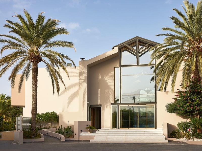 A modern hotel entrance framed by two tall palm trees under a clear blue sky.