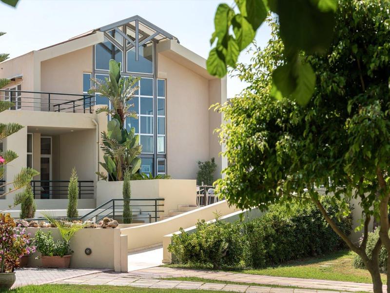 Modern beige hotel building with large windows, balconies, palm trees, and green garden.