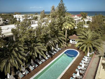 Piscina dell'hotel con lettini e palme, edifici bianchi e mare sullo sfondo.